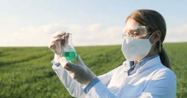 Close up of Caucasian beautiful female ecologist scientist in white gown, mask and googles standing in green field and looking at chemicals in test tube. Woman researcher in margin studying pesticides