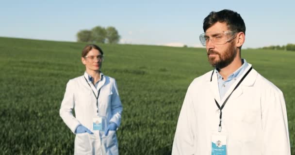 Portrait of Caucasian male ecologist scientist in white gown and googles standing in green field and looking at camera. Female colleague on background. Couple of researchers biologists in margin. — Stock Video