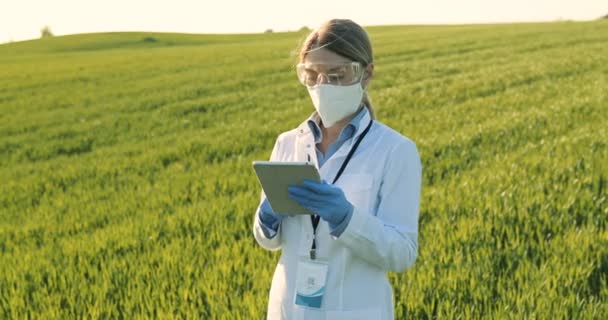 Caucasian beautiful female ecologist scientist in white gown, mask and goggles standing in green field and working on tablet device. Woman researcher, biologist in margin using tablet computer. — ストック動画