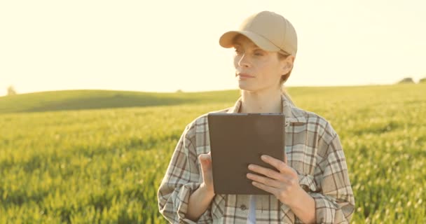 Bonita jovem agricultora de chapéu em pé no campo e usando dispositivo tablet. Mulher muito caucasiana batendo e rolando no computador tablet em margem verde no verão . — Vídeo de Stock