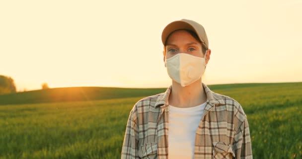 Portrait shot of beautiful young Caucasian woman in mask and hat standing in green field and looking to camera. Close up of female farmer outdoors in summer. Agricultural concept. Pandemic time. — Stock Video