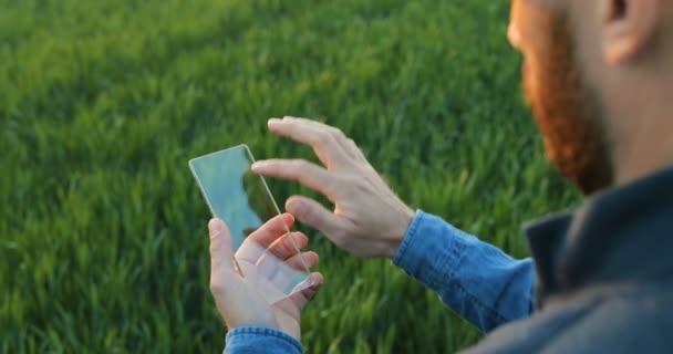 Vue sur l'épaule de l'agriculteur masculin en verre écran transparent dans les mains. Homme tapotant sur dispositif futuriste tout en se tenant dans le champ vert. Écran tactile de haute technologie. Gros plan sur le smartphone du futur.