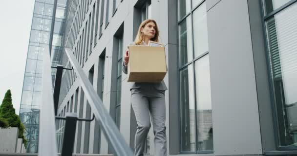 En deprimerad vit kvinnlig kontorsanställd lämnar kontorshuset med en låda med personliga saker. Affärskvinna förlorade jobbet. En eldsjäl som går utomhus. Begreppet "arbetslös utan arbete" — Stockvideo