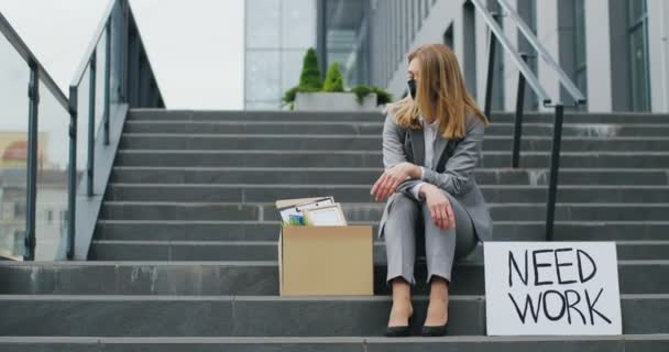 Retrato de una joven desempleada mujer caucásica enmascarada sentada en la escalera al aire libre con póster de cartón Need Work. Desempleo después del bloqueo pandémico. Trabajadora de oficina despedida mirando cosas en la caja . — Vídeos de Stock