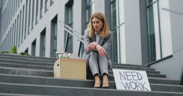 Portret van een blanke werkloze jonge vrouw die buiten op de trap zit met een poster Behoefte aan werk. Vrouwelijke ontslagen werkloos in depressie en angstig op straat met doos van kantoorpersoneel spullen. — Stockvideo