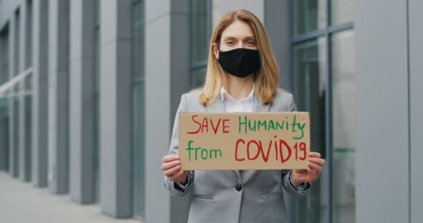 纸盒桌上写着"拯救人类脱离Covid 19"等字。一个戴面具的白人年轻女子的画像，她用焦急的眼睛直视着摄像机。孤独地抗议室外高头牛爆发 — 图库视频影像