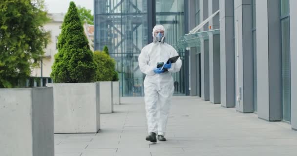 Man i vit kostym, glasögon, handskar och respirator promenader gata i stan. Manlig vårdpersonal med desinficeringsservice som håller mapp och kontaktlös termometer utomhus. Det är pandemisk tid. Desinfektionsmedel — Stockvideo