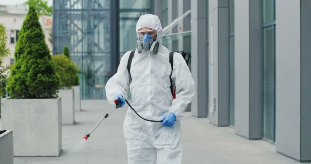 Hombre caucásico en traje blanco antibacteriano especial y respirador caminando por la calle y desinfectando la acera con pulverizador. Pulverización de desinfectante en tierra al aire libre. Concepto pandémico del Coronavirus . — Vídeo de stock