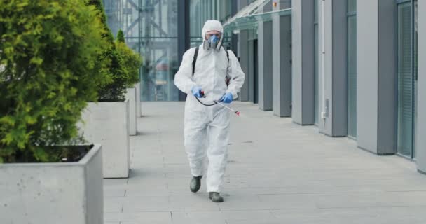 Man in special antibacterial white suit and respirator going by street and disinfecting pavement with pulverizator. Spraying disinfectant on sidewalk outdoors. Covid-19 pandemic outbreak concept. — Stock Video