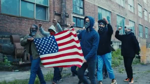 Mixed-races jongens potesters met gemaskerde gezichten die de Amerikaanse vlag vasthouden en in sloppenwijken rondlopen terwijl ze motto 's schreeuwen. Mutie mannelijke rebellen protesteren buiten bij rellen. — Stockvideo