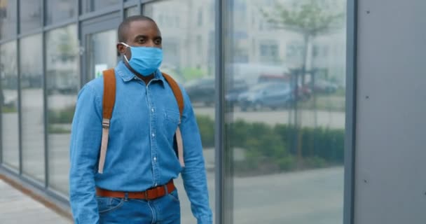Афроамериканец в медицинской маске идёт по улице и куда-то идёт. Мужской пешеход в дыхательной защите прогуливается под открытым небом. Гуляю. Коронавирусная концепция . — стоковое видео