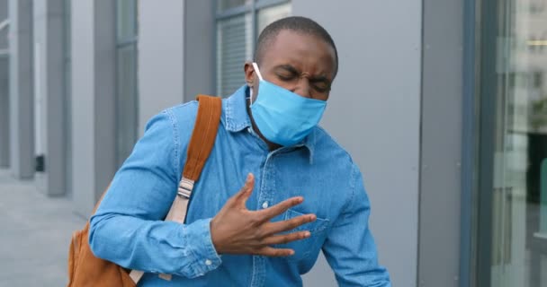 Joven afroamericano con máscara médica caminando por la calle y sintiéndose muy mal. Concepto de enfermedad por Coronavirus. Chico paseando al aire libre, riendo y teniendo dolor en los pulmones . — Vídeos de Stock