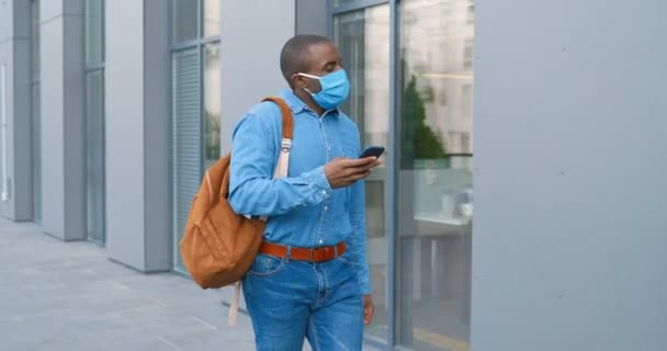 Hombre afroamericano en máscara médica mensaje de texto en el teléfono móvil en la calle. Peatón masculino en protección respiratoria tocando y desplazándose en el teléfono inteligente al aire libre. Chico caminando afuera . — Vídeo de stock