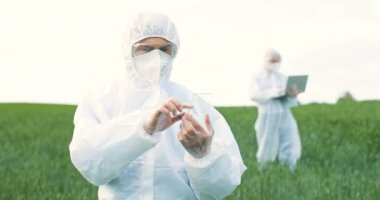 Glass futuistic smartphone in hands of male ecologist standing in green field in summer. Hi-tech technology of transparent device. Man in protective suit tapping on phone of future. Augmented reality.