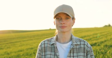 Portrait of pretty young Caucasian woman in hat standing in green field, turning face o camera and smiling cheerfully. Close up of female farmer looking straight with smile outdoors in summer.