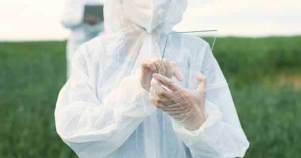 Close up of female farmer ecologist in protective suit tapping on glass transparent screen. Futuristic device in field. Touchscreen of hi-tech. Woman scientist with tablet computer of future in hands. — Stock Video