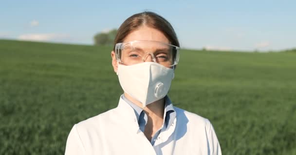 Close up of Caucasian beautiful female ecologist scientist in white gown, mask and googles standing in green field and looking at camera. Portrait shot of woman researcher, biologist in margin. — Stock Video