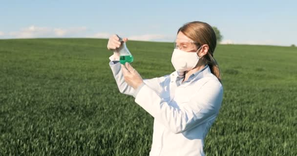 Caucasian pretty female ecologist scientist in white gown, mask and googles standing in green field and looking at chemicals in test tube. Woman researcher and biologist in margin studying pesticides. — Stock Video