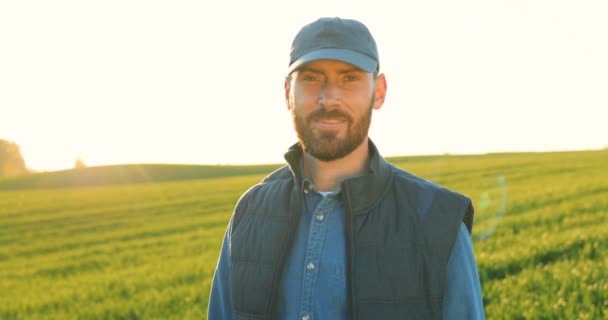 Arckép egy jóképű, fiatal fehér férfiról, kalapban és farmeringben, aki zöld mezőn áll, és boldogan mosolyog a kamerába. Közelkép a férfi farmer szabadban nyáron. A kamera ráközelít a mosolygós arcra — Stock videók