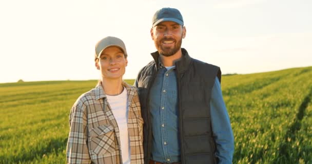 Portret szczęśliwej pary rolników stojących razem na zielonym polu i uśmiechających się radośnie. Ładna kobieta i przystojny mężczyzna przytulający się do upraw pszenicy. Rodzina producentów żywności ekologicznej. — Wideo stockowe