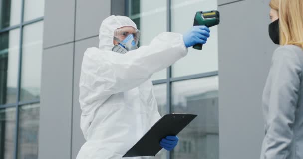 Homme en costume blanc, lunettes et respirateur mesurant la température de la jeune femme avec thermomètre sans contact extérieur et ne laissez pas passer. Un travailleur de la santé trouve un virus chez une piétonne. — Video