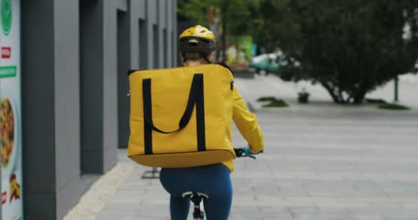 Trasera de mensajería femenina caucásica con gran bolsa de vuelta en casque montar la bicicleta en la calle y la entrega de alimentos o paquetes. Vista posterior de la partera ciclista . — Vídeos de Stock