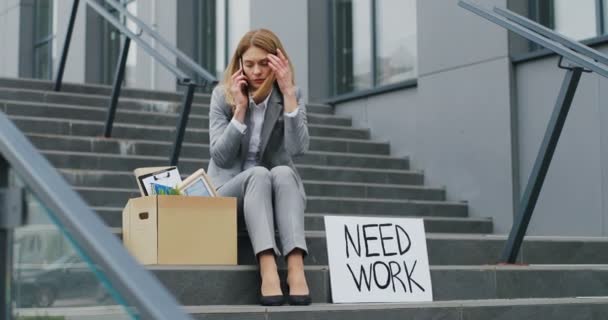 Portrait of young jobless Caucasian woman sitting on stairs outdoor and talking on phone in despair with Need Work board. Unemployment after pandemic lockdown. Workless female speaking on cellphone. — Stock Video