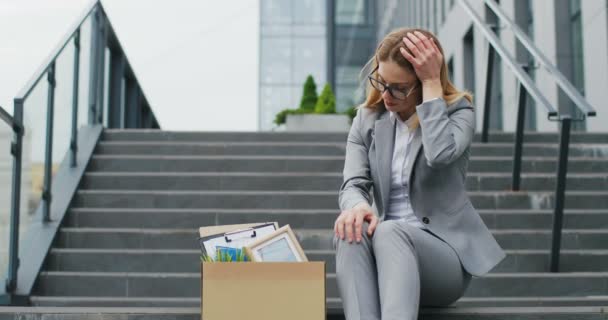 Caucásico joven y triste mujer de negocios sentada en las escaleras al aire libre con caja de cosas como negocio perdido. Trabajadora de oficina desesperada perdió su trabajo. Tasa de desempleo creciente debido a pandemia. Mujer despedida . — Vídeos de Stock