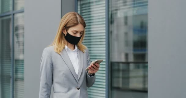Donna d'affari caucasica in maschera che cammina per strada e messaggia sul cellulare. Pedonale femminile in protezione respiratoria passeggiando all'aperto e toccando o scorrendo su smartphone . — Video Stock