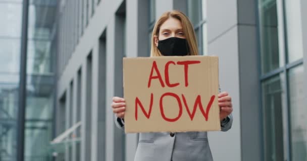 Portrait of Caucasian young woman standing outdoors in town and showing table Act Now. Female demonstrating board with protest against pandemic, political or environmental issues. Single protest. — Stock Video