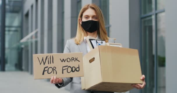 Портрет безробочої жінки в кавказькій масці, що стоїть на відкритому повітрі і показує плакат словами буде працювати на їжу. Звільнена жіноча жартівливість і скринька з офісними речами. — стокове відео