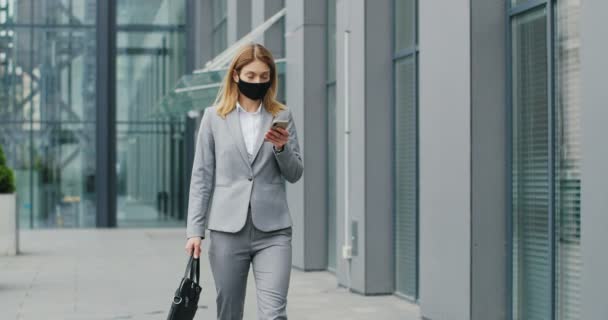 Mujer de negocios caucásica en máscara caminando por la calle y mensajes de texto en el teléfono móvil. Mujer peatonal en protección respiratoria paseando al aire libre y tocando o desplazándose en el teléfono inteligente . — Vídeos de Stock