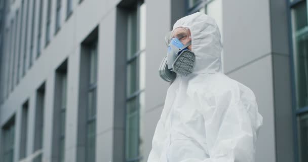 Boční portrét bělocha v bílém obleku, brýle a respirátor čekající na kontrolu teploty chodců. Mužský zdravotnický pracovník dezinfekční služby venku. Koncept pandemické dezinfekce. — Stock video