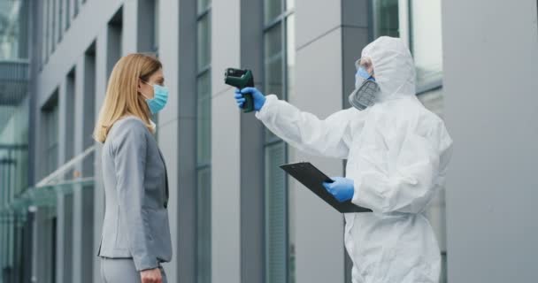 Lavoratrice in maschera che va al lavoro e uomo in completo bianco e respiratore che misura la sua temperatura. Divieto di passaggio al business center per i visitatori. Pandemia di Covid-19. Divieto di ingresso . — Video Stock