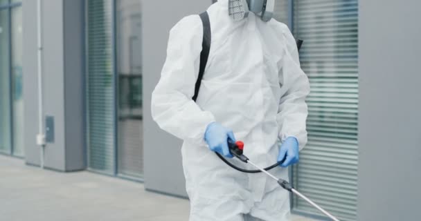 Uomo in speciale abito bianco antibatterico e respiratore che passa per strada e disinfetta pavimentazione con polverizzatore. Spruzzare disinfettante sul marciapiede all'aperto. Concetto di epidemia pandemica Covid-19 . — Video Stock