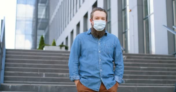 Portrait shot of Caucasian good looking young man with beard and in medical mask standing outdoors, turning face to camera and looking straight. Male pedestrian in respiratry protection at stairs. — Stock Video