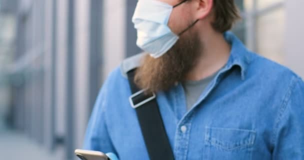 Primer plano del hombre caucásico con barba y en mensaje de texto máscara médica en el teléfono móvil en la calle. Peatón masculino en protección respiratoria tocando y desplazándose en el teléfono inteligente al aire libre . — Vídeo de stock