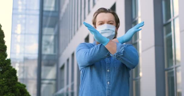 Portret van een jonge blanke man met een medisch masker en handschoenen die op straat staat en geen verboden gebaar met handen toont. Mannelijke kruisarmen zoals stoppen, weigeren of verbieden. Pandemisch concept. — Stockvideo