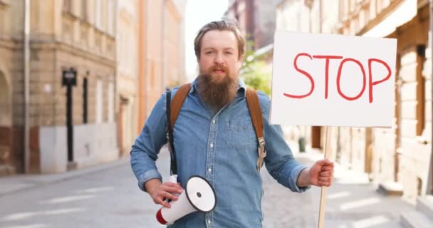 Retrato de un activista caucásico con la barba sosteniendo un afiche Parada y megáfono en una manifestación política o ambiental solitaria en la ciudad. Una sola protesta afuera. Hombre protestando solo . — Vídeo de stock