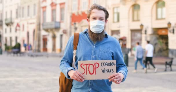 Portret białego aktywisty z brodą i maską medyczną trzymającego plakat Stop Coronavirus na samotnej demonstracji w mieście. Jeden protest na zewnątrz. Koncepcja aktywizmu. Człowiek protestujący samotnie. — Wideo stockowe