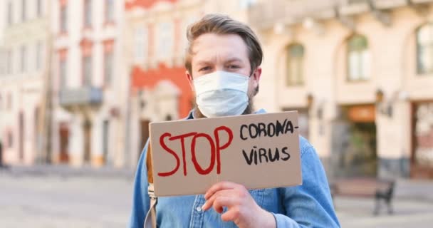 Närbild av kaukasisk man med skägg och i medicinsk mask håller bord Stoppa Coronavirus på en enda protest i staden. En enda protest utanför. Aktivism koncept. Porträtt av manliga protesterande ensam. — Stockvideo