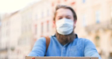 Karton masayı kapatın ve insanlığı Covid 19 'un elinden tıbbi maskeli beyaz bir gencin elinden kurtarın. Arkasında bulanık erkek eylemci var. Dışarıda yapayalnız protesto. Sağlık hizmetleri aktivizmi.