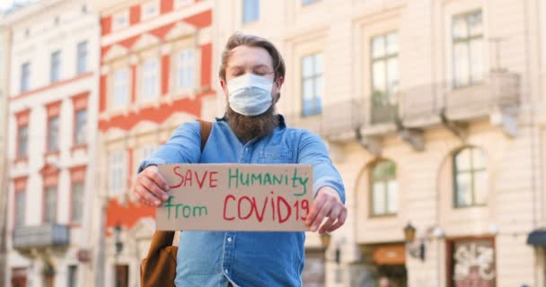 Porträtt av kartong affisch med ord Rädda mänskligheten från Covid 19 i händerna på vit ung man i medicinsk mask. Ensam protestera utomhus på torget. Manlig hälsovårdsaktivist. — Stockvideo