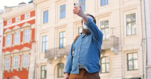 Homme caucasien avec barbe et masque médical debout dans la rue et vidéochat sur téléphone mobile. Homme en protection respiratoire est à l'extérieur tout en parlant et en ayant vidéochat par webcam sur smartphone — Video