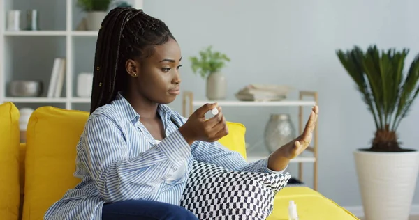 สาวแอฟริกันอเมริกันที่น่ารักนั่งบนโซฟาในห้องนั่งเล่นและขัดเล็บของเธอ ในร่ม — ภาพถ่ายสต็อก