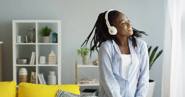 Kaunis nuori iloinen afroamerikkalainen nainen tanssii musiikin tahdissa, jota hän kuuntelee kuulokkeilla. Kotona. Sisällä — kuvapankkivalokuva