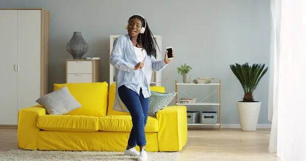 Lykkelig ung afroamerikansk kvinne i jeans og skjorte som danser fint i stua mens hun hører på musikk på smarttelefonen. Innendørs – stockfoto