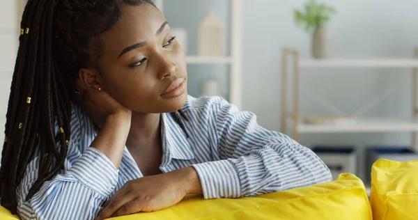 Viehättävä afroamerikkalainen nuori nainen sinisessä paidassa istuu keltaisella sohvalla ja miettii ja hymyilee kameralle. Sisätilat — kuvapankkivalokuva