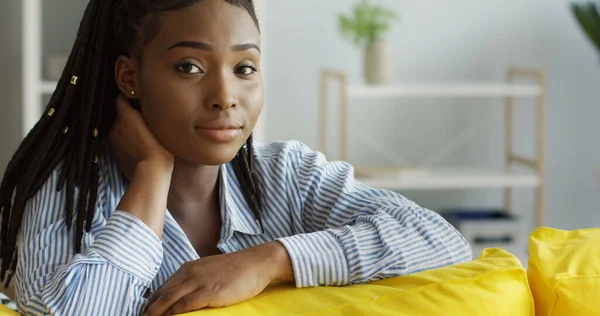 Sjarmerende afroamerikansk ung kvinne i blå skjorte sitter på den gule sofaen og tenker, og smiler så til kameraet. Innendørs – stockfoto