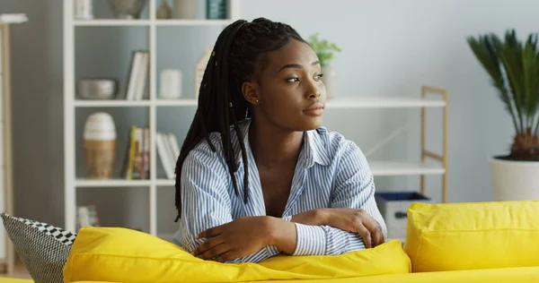 Retrato da jovem mulher afro-americana atraente com uma cauda de tranças apoiada no sofá amarelo na sala de estar, olhando para o lado e depois sorrindo direto para a câmera. Para dentro — Fotografia de Stock